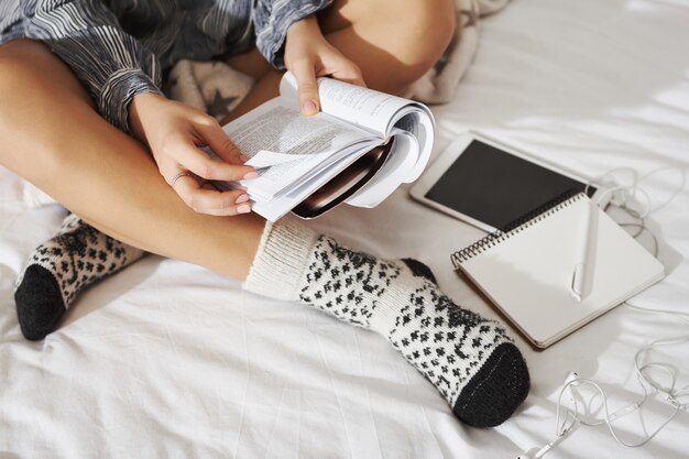 Tiro de ángulo lateral de la mujer sentada con las manos cruzadas en la cama, con calcetines elegantes, tomando notas mientras estudiaba en casa. Alumno trabajando en la tarea, usando tableta digital, escuchando música con auriculares