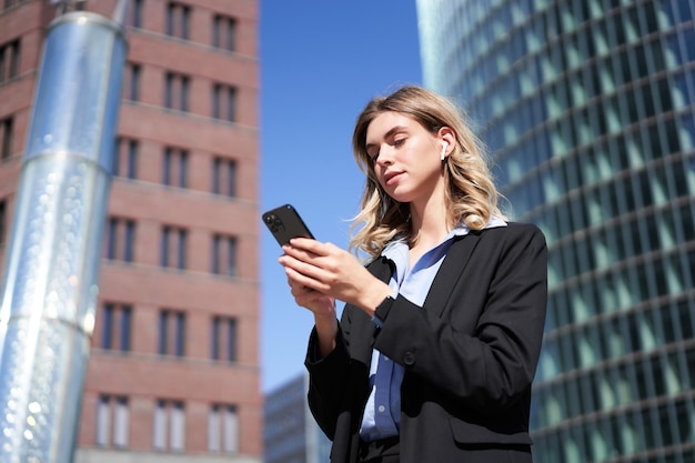 Foto gratuita tiro de ángulo bajo de mujer joven gerente empresaria en traje sosteniendo teléfono móvil de pie con smar