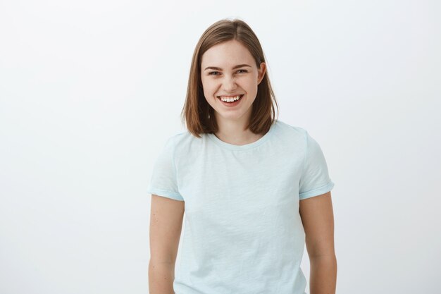 Tiro de cintura para arriba de alegre, entretenida y divertida, guapa morena ordinaria en camiseta de moda riendo felizmente y mirando tener una divertida charla divertida con amigos sobre la pared gris