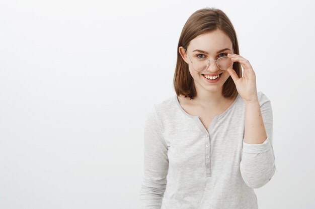 Tiro de cintura para arriba de alegre mujer creativa e inteligente con cabello castaño corto mirando desde debajo de la frente misteriosa e intrigada quitándose las gafas y sonriendo ampliamente divirtiéndose sobre la pared gris