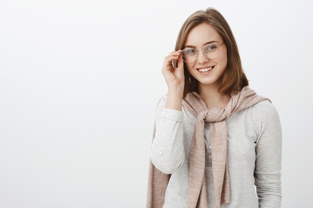 Tiro de cintura para arriba de atractiva mujer joven con estilo con gafas y jersey rosa atado sobre el cuello con una blusa tocando anteojos y sonriendo amistosamente pasando el rato en compañía desconocida