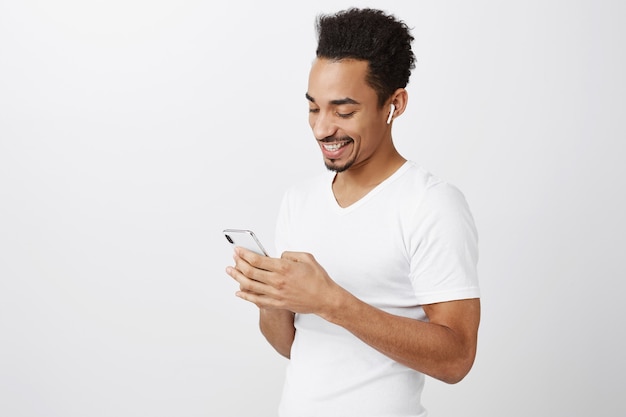 Tiro de cintura para arriba de atractivo hombre afroamericano charlando, enviando mensajes de texto a un amigo, escuchando música o viendo videos en auriculares inalámbricos