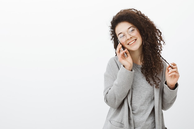 Tiro de cintura para arriba de una chica elegante y amigable saliente con cabello rizado en elegantes gafas y abrigo gris, hablando por teléfono inteligente, inclinando la cabeza y sonriendo ampliamente