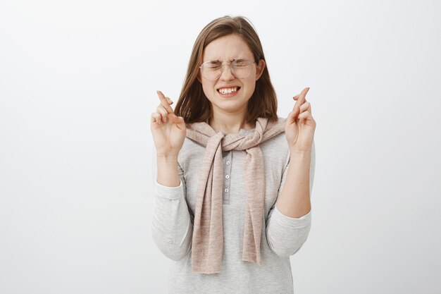 Tiro de cintura para arriba de una chica inteligente, guapa y nerviosa en blusa y suéter atado sobre el cuello, cerrando los ojos y apretando los dientes mientras se siente intensamente cruzando los dedos por la buena suerte pidiendo deseos sobre una pared gris