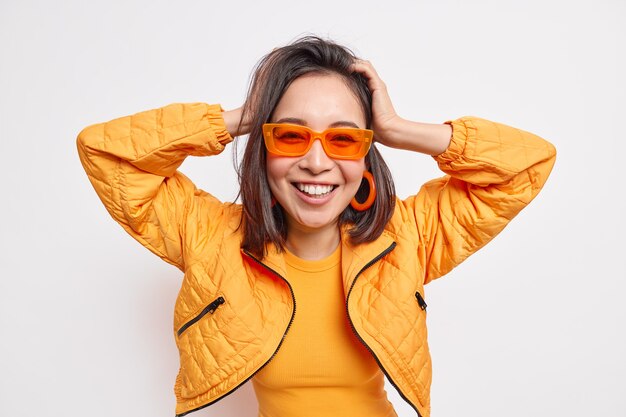 Tiro de cintura para arriba de la chica milenaria asiática de moda positiva tiene el pelo oscuro, mantiene las manos en la cabeza, sonríe ampliamente, usa una chaqueta de gafas de sol de moda aislada sobre una pared blanca de buen humor.