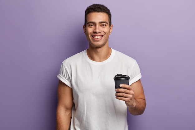 Tiro de cintura para arriba de un chico guapo alegre y sonriente sostiene café de papel para llevar, disfruta del tiempo libre con un amigo, bebe bebidas aromáticas, viste una camiseta blanca, modelos contra la pared púrpura