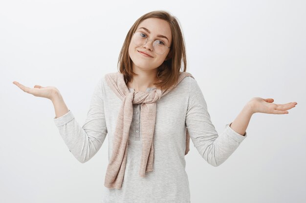 Tiro de cintura para arriba de desorientada despreocupada atractiva chica europea con gafas encogiéndose de hombros con las manos extendidas a un lado sonriendo inclinando la cabeza sin tener ni idea de ser inconsciente y descuidado sobre la pared gris