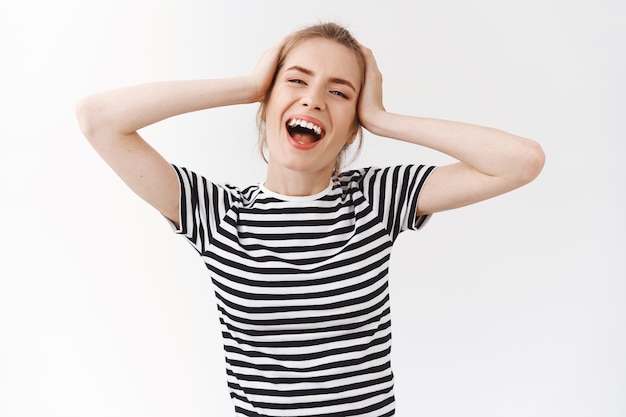 Tiro de cintura para arriba, despreocupada, vivaz mujer joven emotiva con moño desordenado en camiseta a rayas, sonriendo cantando y disfrutando de un hermoso día, sintiéndose relajado y encantado, tocando la cabeza bailando, fondo blanco