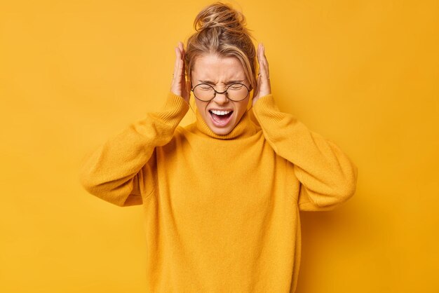 Tiro de cintura arriba de emocional joven mujer rubia grita en voz alta cubre los oídos con las manos no quiere escuchar un sonido molesto usa gafas y suéter aislado sobre fondo amarillo