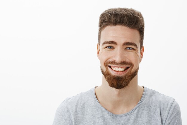 Tiro de cintura para arriba del encantador hombre guapo despreocupado y optimista con barba, bigote y lindos ojos azules riendo alegremente entrecerrando los ojos de alegría y diversión divirtiéndose contra la pared gris