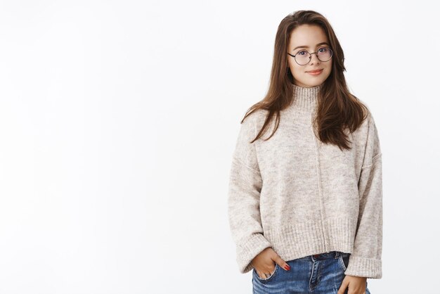 Tiro de cintura para arriba de la encantadora estudiante europea con linda sonrisa en gafas y suéter cálido acogedor sosteniendo la mano en el bolsillo y mirando interesado y despreocupado en gris.