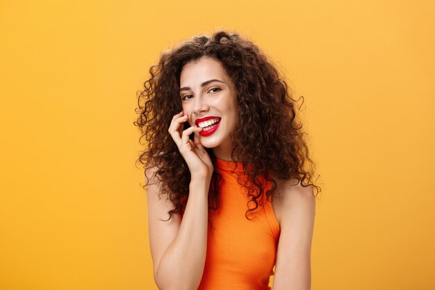 Tiro de cintura para arriba de la encantadora mujer femenina tonta y despreocupada con peinado rizado en la parte superior recortada mordiendo el dedo y sonriendo sensual y coqueta a la cámara con un aspecto sexy y atrevido sobre fondo naranja.