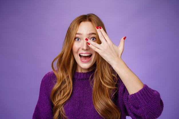 Tiro de cintura para arriba de entusiasta y carismática mujer pelirroja divertida linda en suéter púrpura con uñas frescas que muestran asomándose a través de agujeros en los dedos y sonriendo optimista y emocionada, mirando sorprendido.