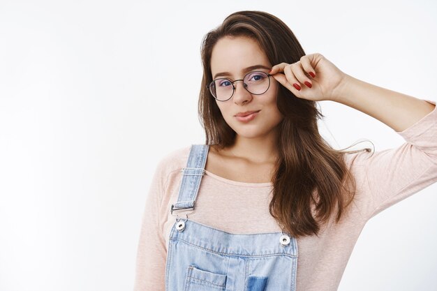 Tiro de cintura para arriba de la estudiante atractiva sensual y coqueta con el pelo castaño que toca el marco de los vidrios que doblan los labios lindos que hacen miradas románticas al frente que se colocan seductoras sobre la pared gris.