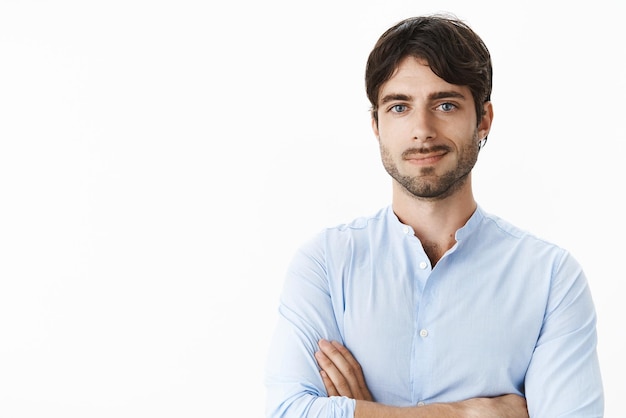 Foto gratuita tiro de cintura para arriba del exitoso hombre joven apuesto encantado con ojos azules y barba sonriendo satisfecho mientras se toman de las manos cruzadas sobre el pecho siendo autosatisfecho logrando buenos ingresos en la pared gris.