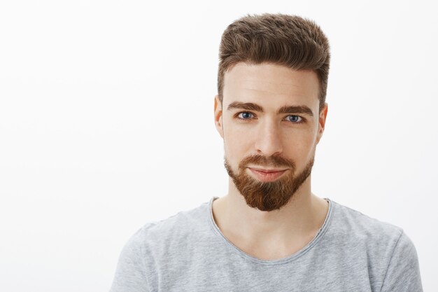 Tiro de cintura para arriba de guapo sensual y confiado joven con barba, bigote y ojos azules sonriendo mirando sincero y seguro de sí mismo como con una sonrisa encantada contra la pared gris