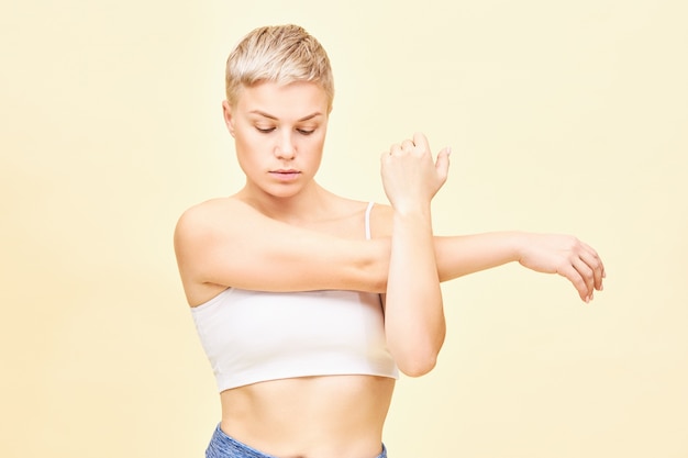 Tiro de cintura para arriba de hermosa mujer joven de moda con corte de pelo juvenil con mirada concentrada, centrada en sus sentimientos mientras estira los músculos del brazo, presionando el hombro y el codo contra su pecho