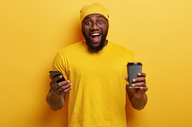 Tiro de cintura para arriba de hipster étnico feliz desarrolla un sitio web personal en un teléfono celular, conectado a internet inalámbrico, sostiene una taza desechable de bebida caliente, tiene una barba gruesa, usa un sombrero amarillo y una camiseta