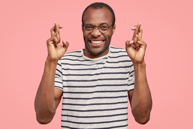 Tiro de cintura para arriba de hombre alegre de piel oscura tiene una sonrisa con dientes, cruza los dedos para la buena suerte