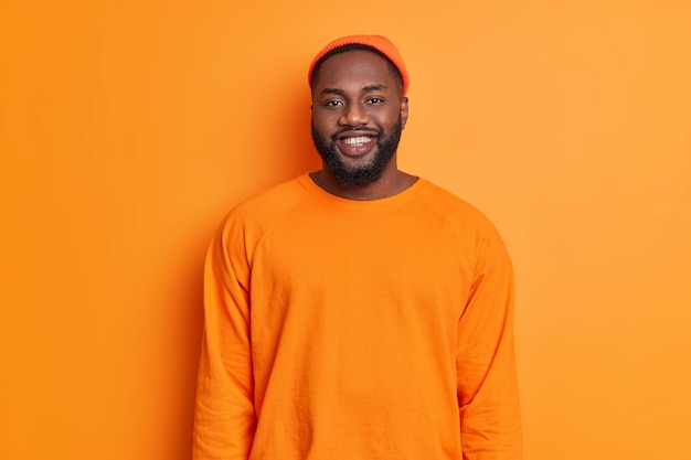 Tiro de cintura para arriba de hombre feliz sonríe felizmente vestido con sombrero naranja y suéter de buen humor mira directamente al frente expresa emociones positivas se encuentra en estudio contra una pared brillante