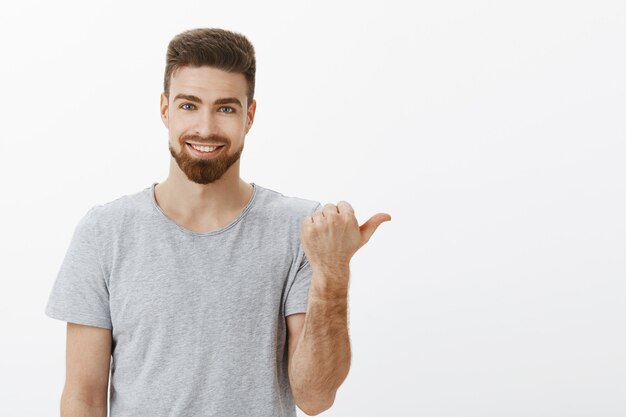 Tiro de cintura para arriba de hombre guapo, elegante y masculino seguro de sí mismo con barba y linda sonrisa blanca apuntando a la derecha con el pulgar asegurando que el espacio de la copia es perfecto con una expresión de confianza y alegría