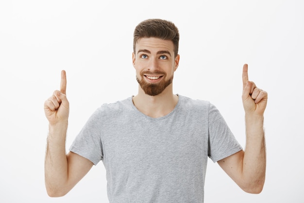 Tiro de cintura para arriba de hombre guapo masculino con ojos azules y barba sonriendo alegremente y seguro de sí mismo levantando las manos mirando y apuntando hacia arriba encantado y feliz de pie complacido sobre la pared gris