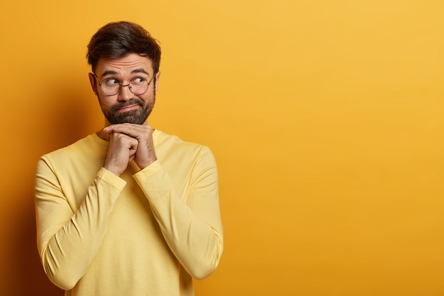 Tiro de cintura para arriba de un joven barbudo que piensa seriamente en su oferta, mantiene las manos debajo de la barbilla, se concentra a un lado, usa lentes ópticos y un suéter, sueña con algo, aislado en una pared amarilla