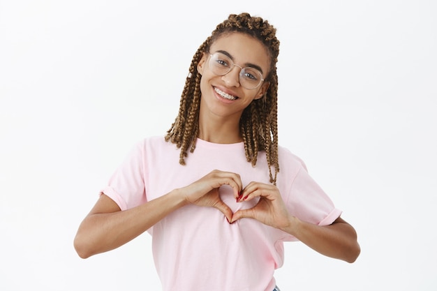 Tiro de cintura para arriba de linda novia afroamericana atractiva y amigable con rastas en camiseta rosa mostrando gesto de corazón