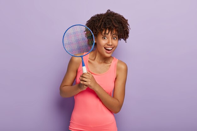 Tiro de cintura para arriba de la mujer afroamericana encantada sostiene la raqueta de tenis, espera a que un amigo tenga el torneo