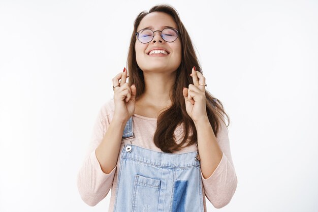 Tiro de cintura para arriba de una mujer atractiva soñadora esperanzada con gafas levantando la cabeza en el cielo con los ojos cerrados y una sonrisa feliz y optimista mientras ora pidiendo a Dios que haga realidad el sueño, cruce los dedos para la buena suerte.