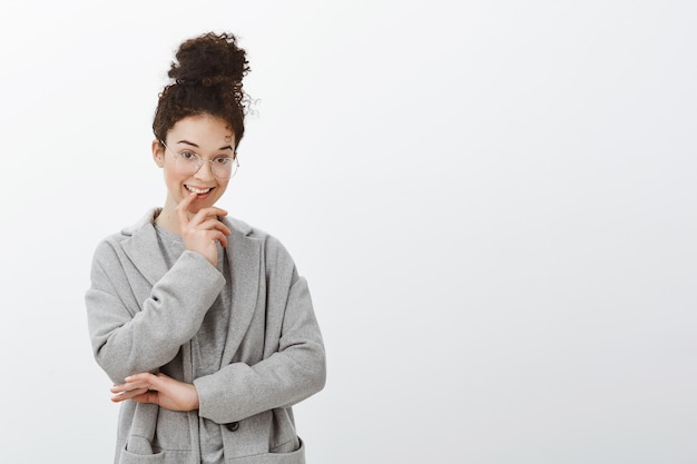 Tiro de cintura para arriba de mujer creativa de pelo rizado inteligente con peinado moño con gafas de moda y abrigo gris, mordiendo el dedo y sonriendo con expresión intrigada