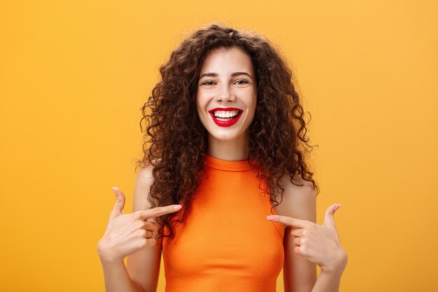 Tiro de cintura para arriba de mujer encantadora saliente orgullosa y feliz con cabello rizado y lápiz labial rojo apuntando a sí misma encantada y confiada hablando de sus propios logros sobre fondo naranja. Copia espacio