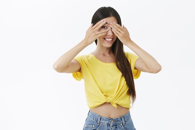 Foto gratuita tiro de cintura para arriba de la mujer europea linda intrigada y alegre creativa en la camiseta amarilla de moda que cubre los ojos con las palmas