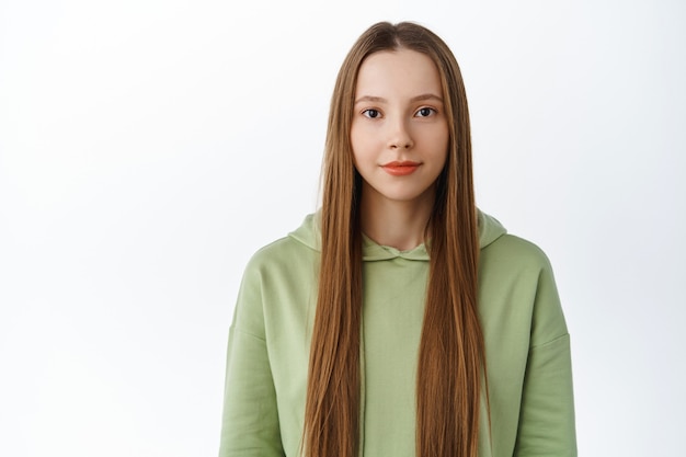 Tiro de cintura para arriba de mujer hermosa joven con cabello largo sano, maquillaje desnudo natural, sonriendo y mirando determinado al frente, de pie en sudadera con capucha contra la pared blanca