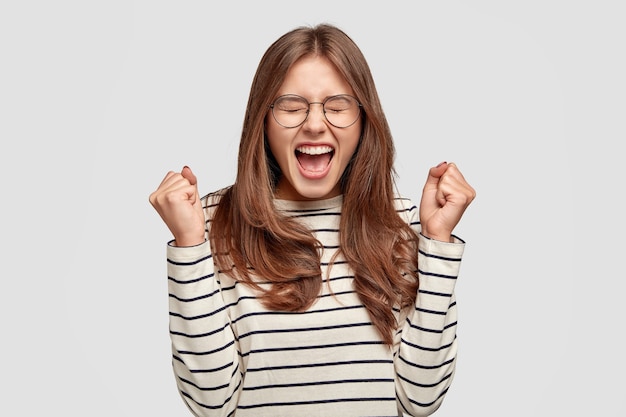 Tiro de la cintura para arriba de la mujer joven autodeterminada y llena de alegría levanta las manos con los puños cerrados, aclama el éxito