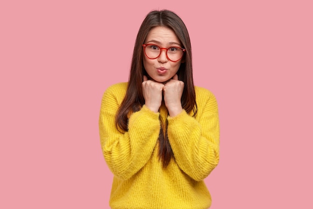 Tiro de cintura para arriba de mujer joven y bonita mantiene las manos en puños debajo de la barbilla, tiene los labios doblados, usa gafas transparentes, suéter amarillo casual