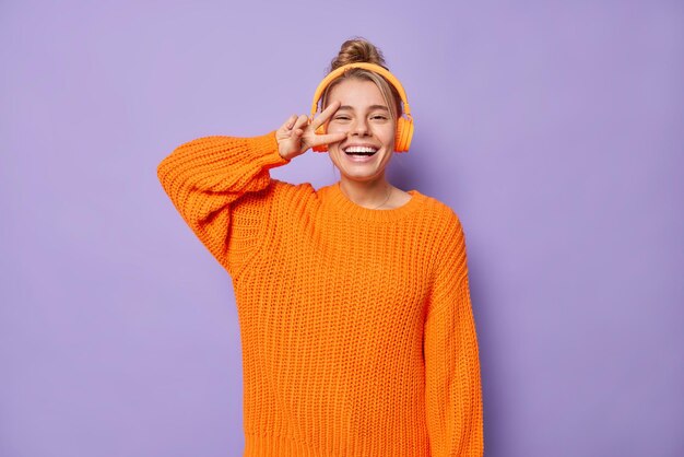 Tiro de cintura arriba de una mujer joven positiva y alegre con un moño de pelo sonríe con dientes hace un gesto de paz sobre el ojo se divierte escucha música a través de auriculares estéreo posa contra un fondo púrpura Lenguaje corporal