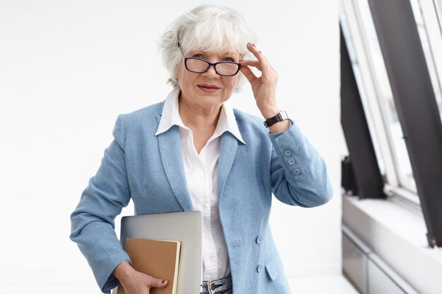 Tiro de la cintura para arriba de la mujer madura de pelo gris de mediana edad con elegante chaqueta azul y camisa blanca ajustando sus anteojos, posando en el interior de la oficina, llevando una computadora portátil y un diario en su camino a la reunión