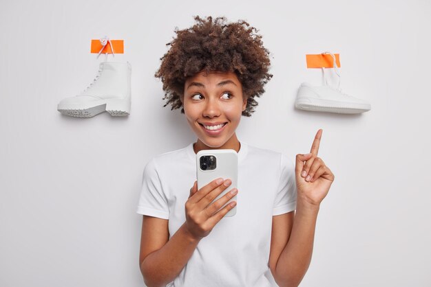 Tiro de cintura para arriba de una mujer morena alegre que señala con el dedo índice arriba en los zapatos enlucidos sonríe con dientes recomienda algo para usar usa el teléfono móvil navega en la tienda en línea aislada sobre la pared blanca del estudio