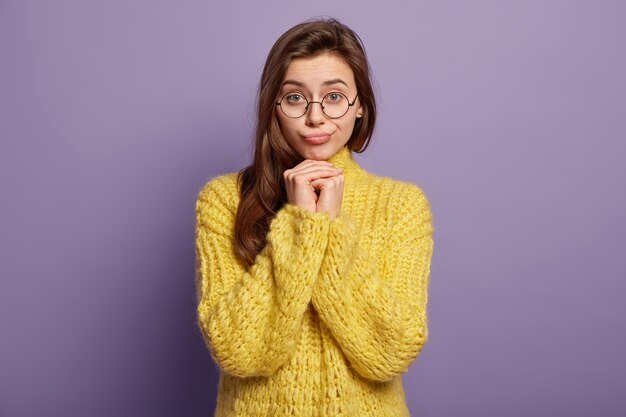 Tiro de cintura para arriba de una mujer morena bonita que se siente sola y triste, mantiene las manos debajo de la barbilla, molesta por las malas noticias, usa un jersey amarillo, se para sobre una pared púrpura. Expresiones y sentimientos faciales humanos