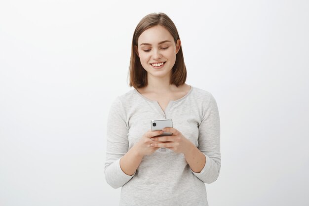 Tiro de cintura para arriba de la mujer morena europea ordinaria moderna en traje casual con teléfono inteligente sonriendo en la pantalla del dispositivo enviando mensajes de texto o editando fotos para publicar en línea posando sobre una pared gris