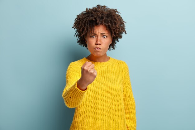 Tiro de cintura para arriba de mujer de piel oscura insatisfecha hosca tiene peinado Afro, agita el puño y mira con amenaza