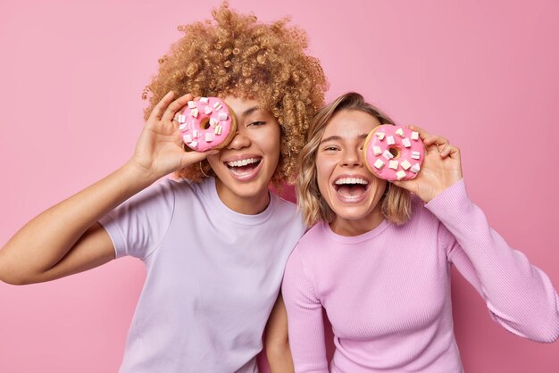 Tiro de cintura arriba de mujeres jóvenes y amigables que se cubren los ojos con deliciosos donuts, diviértanse, disfruten de comer postres dulces, estén cerca unos de otros, aislados sobre un fondo rosa. Donuts sabrosos