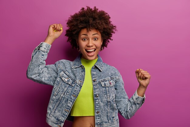 Tiro de cintura para arriba de optimista alegre mujer afroamericana levanta la mano, se siente optimista, se mueve felizmente, aprieta los puños, usa chaqueta de mezclilla, aislada sobre una pared vibrante púrpura. Concepto de estilo de vida feliz