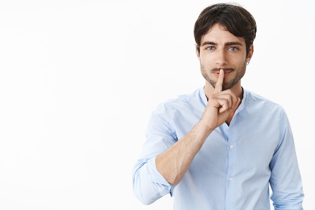 Tiro de cintura para arriba de sexy apuesto hombre exitoso con barba y ojos azules sonriendo travieso haciendo gesto de silencio sobre los labios doblados pidiendo mantener la voz baja mientras prepara una sorpresa secreta sobre la pared gris