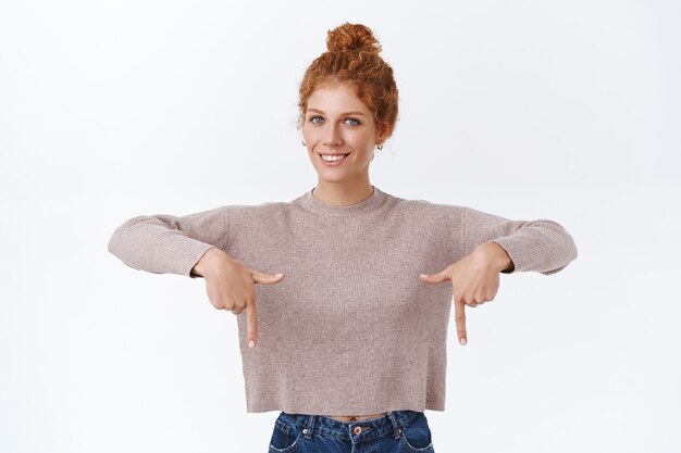 Tiro de cintura para arriba tierna, encantadora mujer caucásica pelirroja con cabello rizado peinado en un moño, usa un suéter cálido y acogedor, apuntando hacia abajo y sonriendo feliz cámara, publicitar promocional, dar consejos, pared blanca