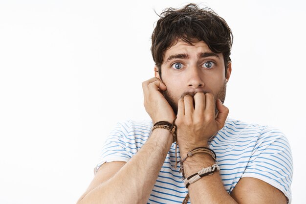 Tiro de cintura para arriba de tímido e inseguro chico guapo joven tonto con ojos azules innosent mordiendo las uñas sosteniendo la mano en la mejilla sintiendo pánico y preocupación de pie ansioso y asustado sobre la pared gris