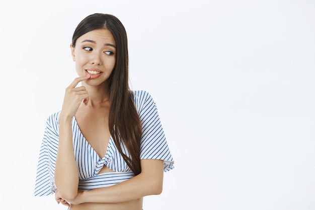 Foto gratuita tiro de cintura para arriba de tonta mujer asiática femenina y linda con cabello oscuro en blusa rayada recortada mordiendo el dedo mirando suavemente la esquina inferior derecha