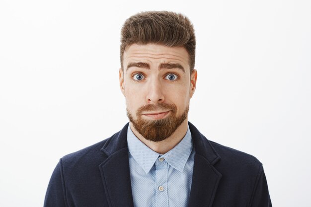 Tiro de cintura para arriba del tonto y sombrío novio moreno lindo con ojos azules y barba sonriendo de lado de pie en traje elegante sintiéndose arrepentido y disculpándose por ser estúpido posando contra la pared gris