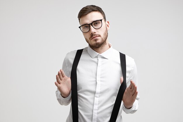 Tiro de cintura para arriba del trabajador de oficina masculino joven confiado de moda con barba y corte de pelo de moda de pie en la pared blanca con gafas elegantes y camisa formal, tirando de sus tirantes negros